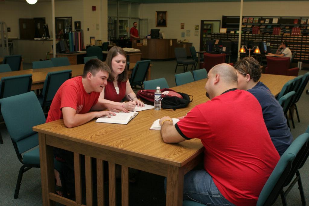 Four people are seated around a wooden table in a library, engaged in discussion. One man, holding a pen, points to a notebook. Others listen attentively. Shelves and desks are visible in the background.