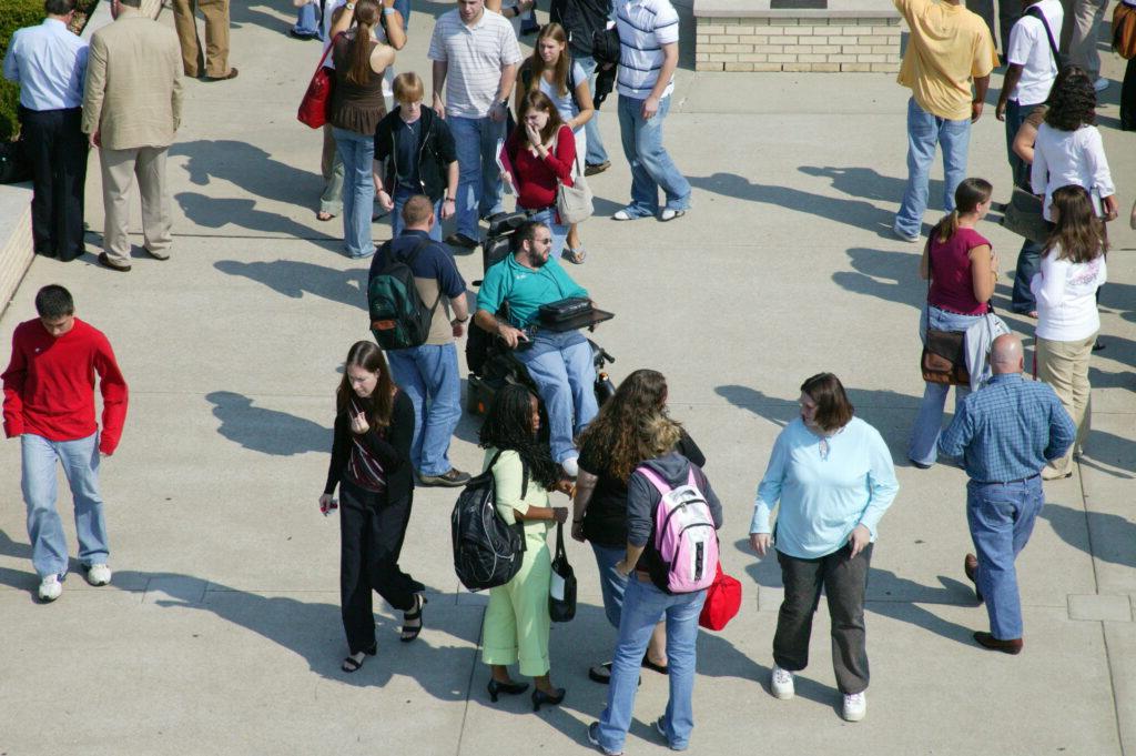 A diverse group of people, including students with backpacks and a person in a wheelchair, are mingling and walking in an outdoor paved area surrounded by buildings.